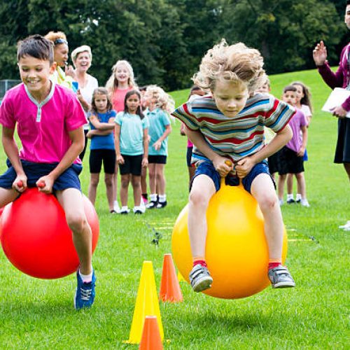 Little boys sitting on  space hoppers racing,  classmates stand in the background watching during a sports lesson.